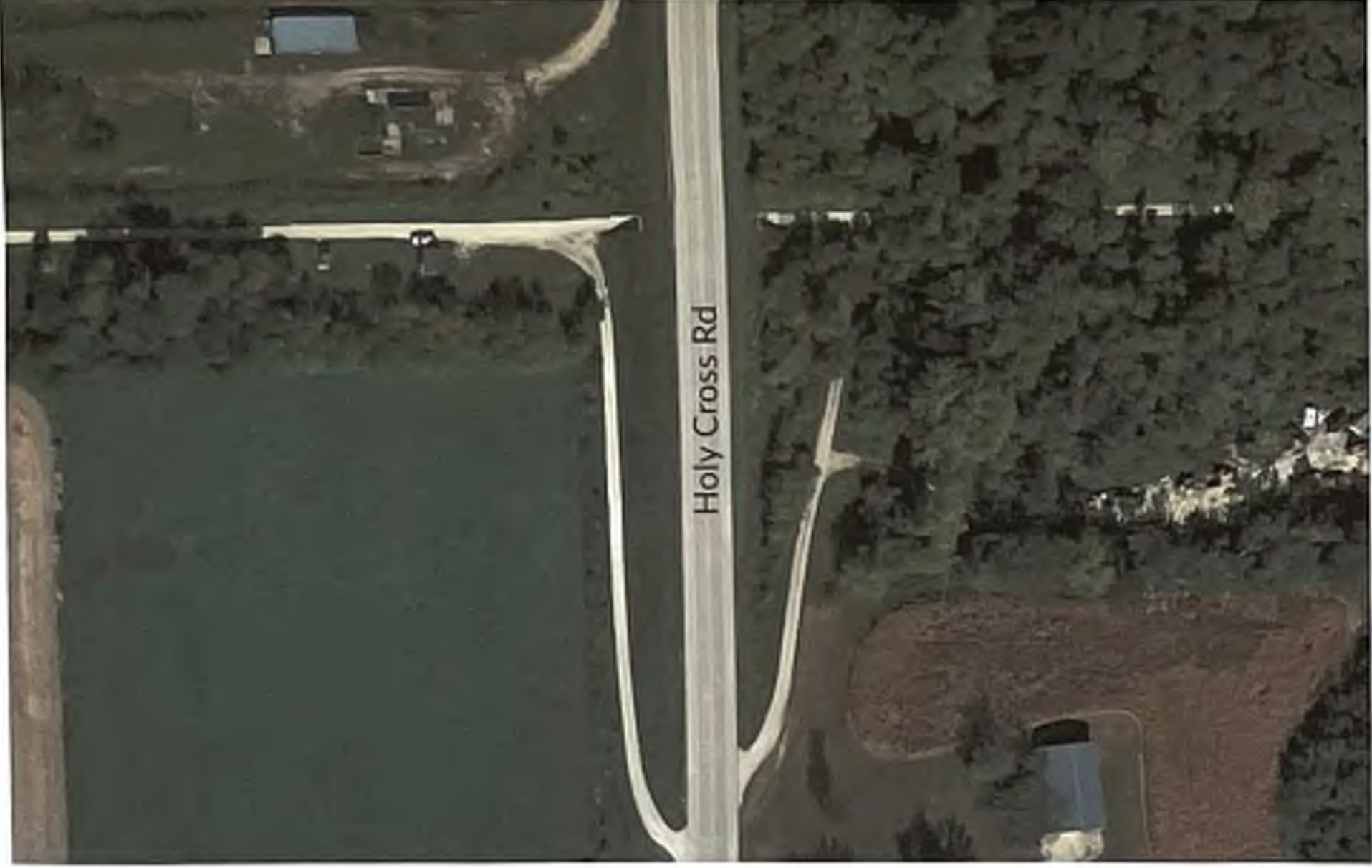 The map displays an aerial view of Holy Cross Rd, surrounded by agricultural fields and some buildings. The road extends through a rural area, with forested regions adjacent to the east and notable landmarks like a blue roof structure visible in the lower section of the image. This layout provides insight into the spatial relationships in the vicinity of Holy Cross Rd.
