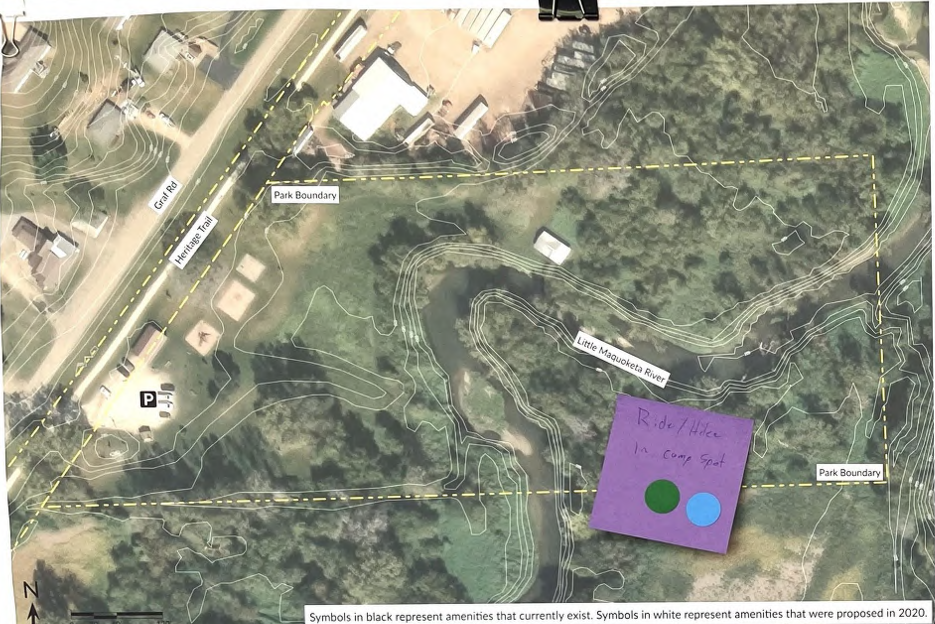 This map displays the layout of a park, including labeled trails such as Heritage Trail, the Park Boundary outlined in yellow, parking areas marked with a 'P', and the Little Maquoketa River forming part of the landscape. Symbols indicate existing amenities in black and proposed amenities in white, providing a visual reference for park features and accessibility routes.