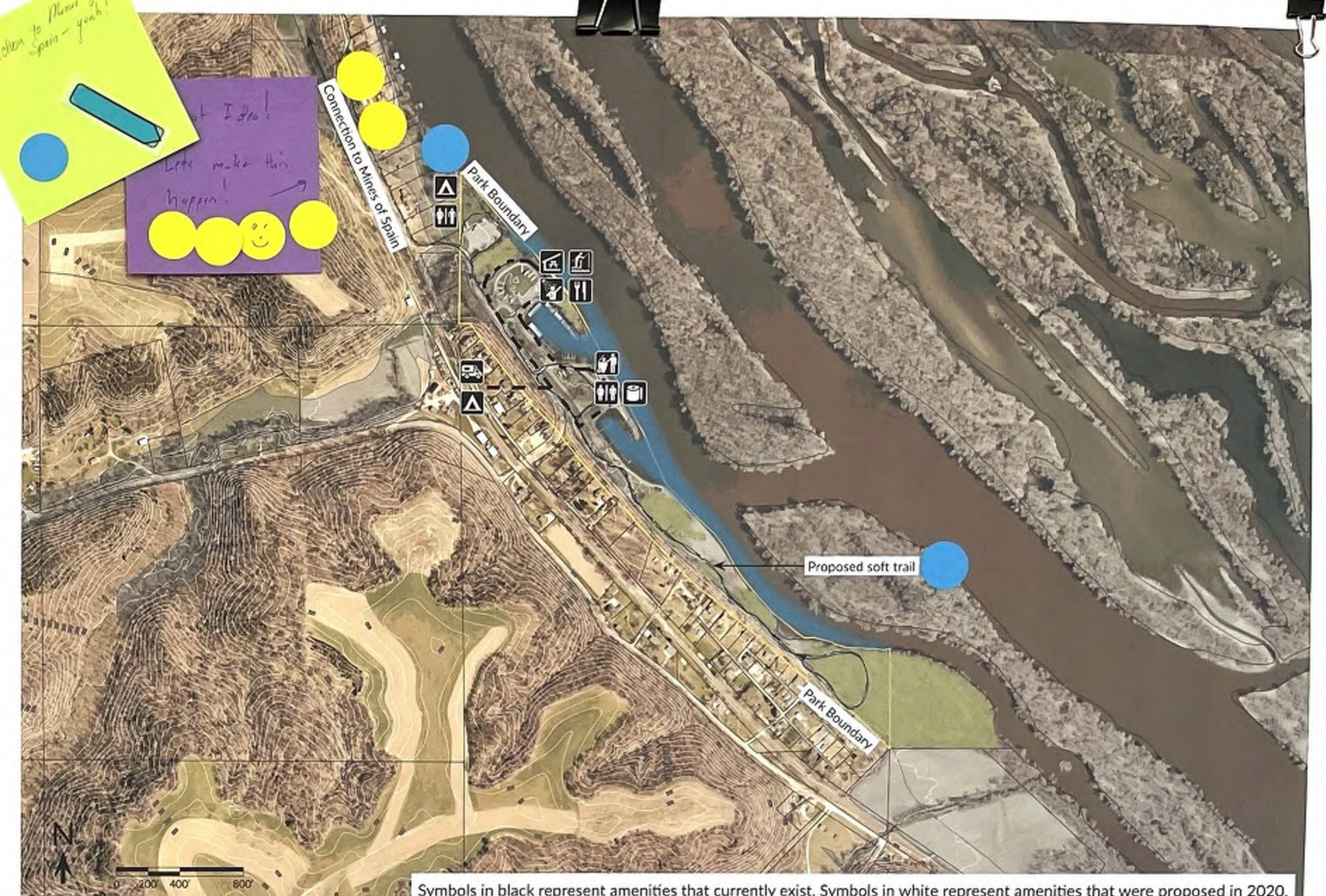 The map depicts a river area with existing amenities indicated by black symbols and proposed amenities shown by white symbols, including a proposed soft trail labeled on the map. Colorful note stickers are placed around the map with text messages that suggest planning and brainstorming activities. The spatial layout includes the river, nearby land features, and the concept design for future developments in the area.