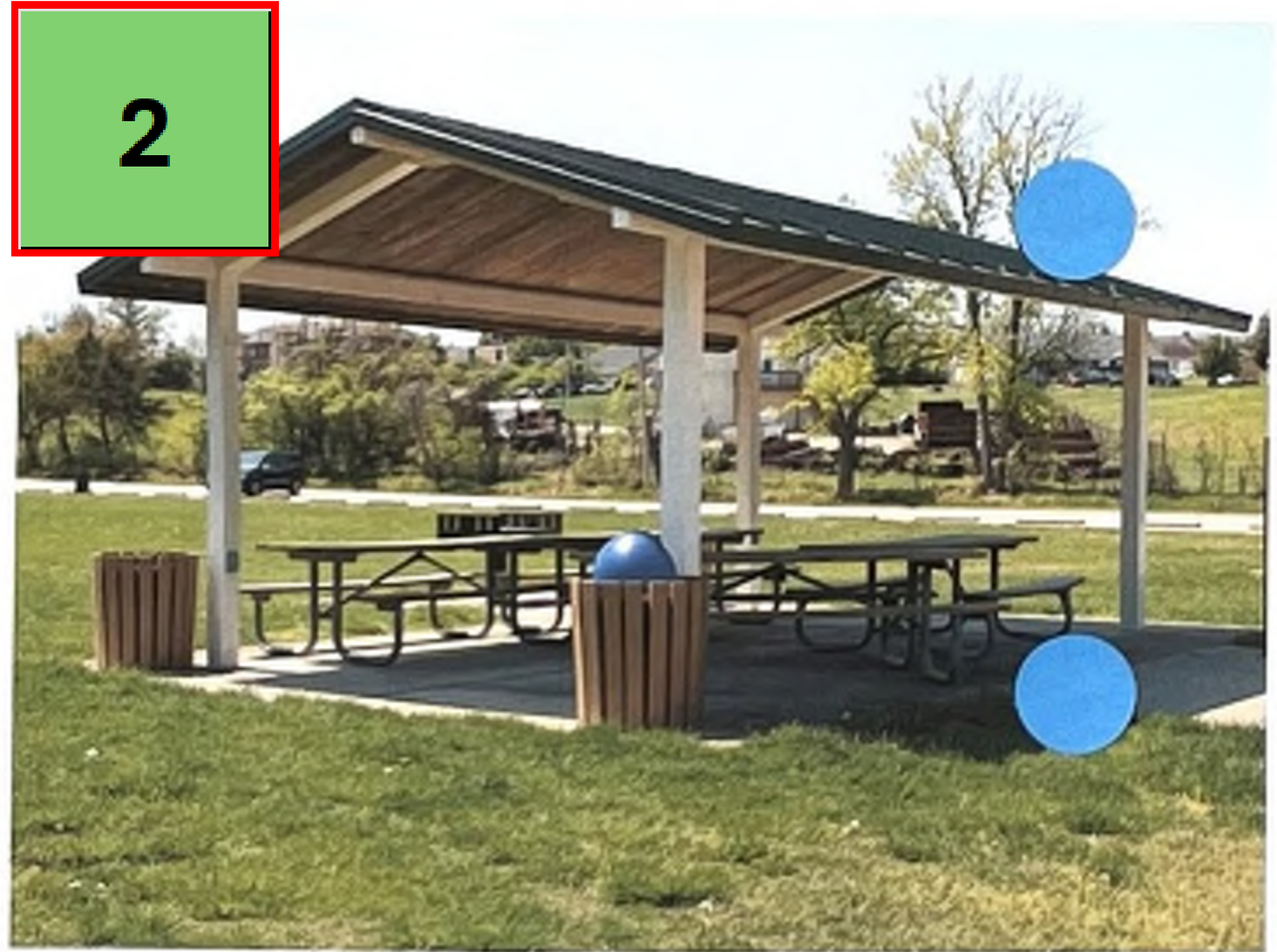 The image depicts a picnic pavilion situated in a grassy park area. The pavilion has a wooden roof supported by four columns and features multiple picnic tables underneath. Two blue circles are placed on the image, indicating specific points of interest. The background shows trees and a parking area, providing context to the location.