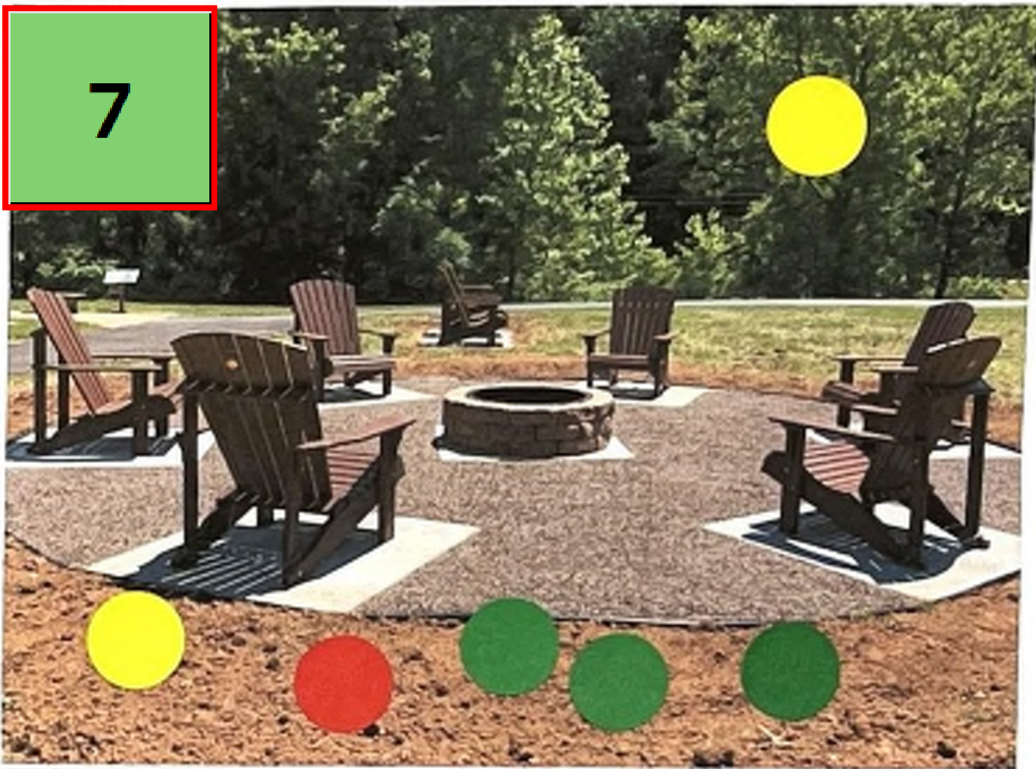 The image shows an outdoor seating area featuring several wooden Adirondack chairs positioned around a central fire pit, which is made of stone. The seating area is set on a gravel-like surface with concrete pads below each chair. Around the seating area, there are green trees and grassy areas. There is a numbered sign in a green box on the left side of the image, and colored circles at the bottom which may represent some kind of key or legend.