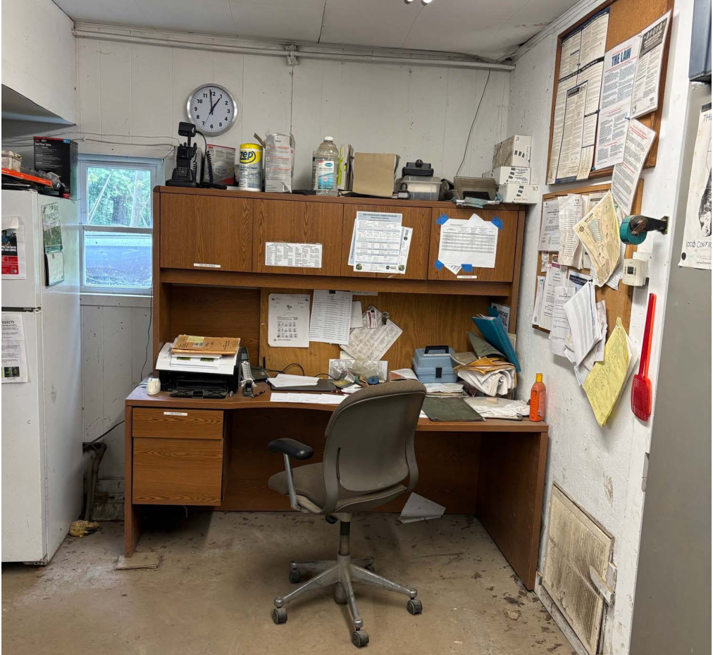 The image depicts a cluttered office environment that includes a wooden desk with an office chair, a printer on the desk, and several papers scattered across the work surface. Above the desk is a wall-mounted clock and a bulletin board with multiple documents pinned to it. A window is visible on the left side, and a refrigerator is located on the far left. The floor appears to be bare concrete, adding to the utilitarian look of the space.