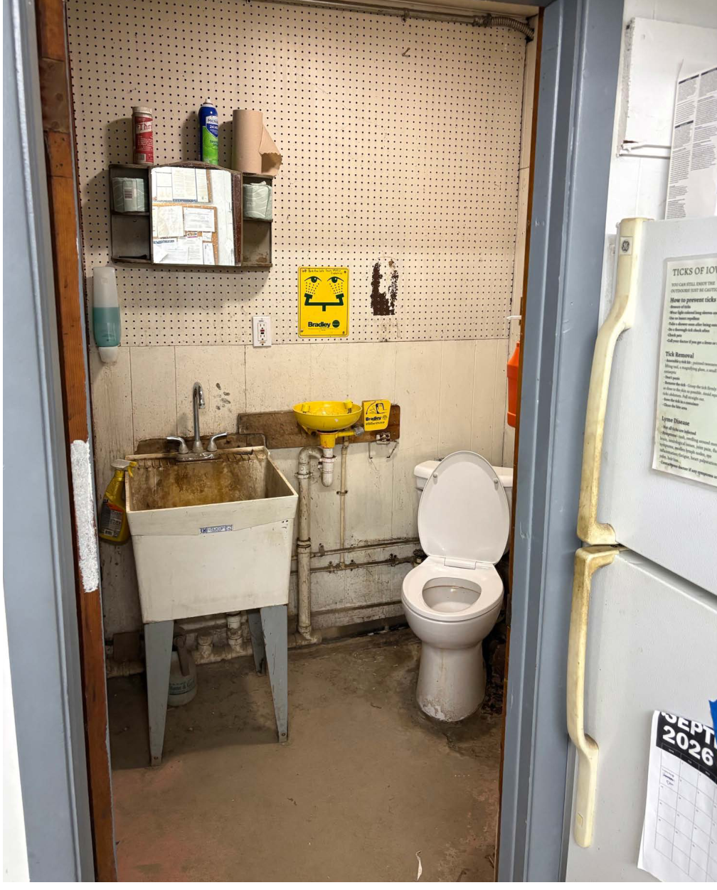 The image shows a small restroom featuring a white toilet and a dirty sink. On the wall are several shelves with cleaning supplies, a mirror, and a yellow safety sign indicating eye wash station. The flooring is concrete, and the overall environment appears utilitarian and functional.