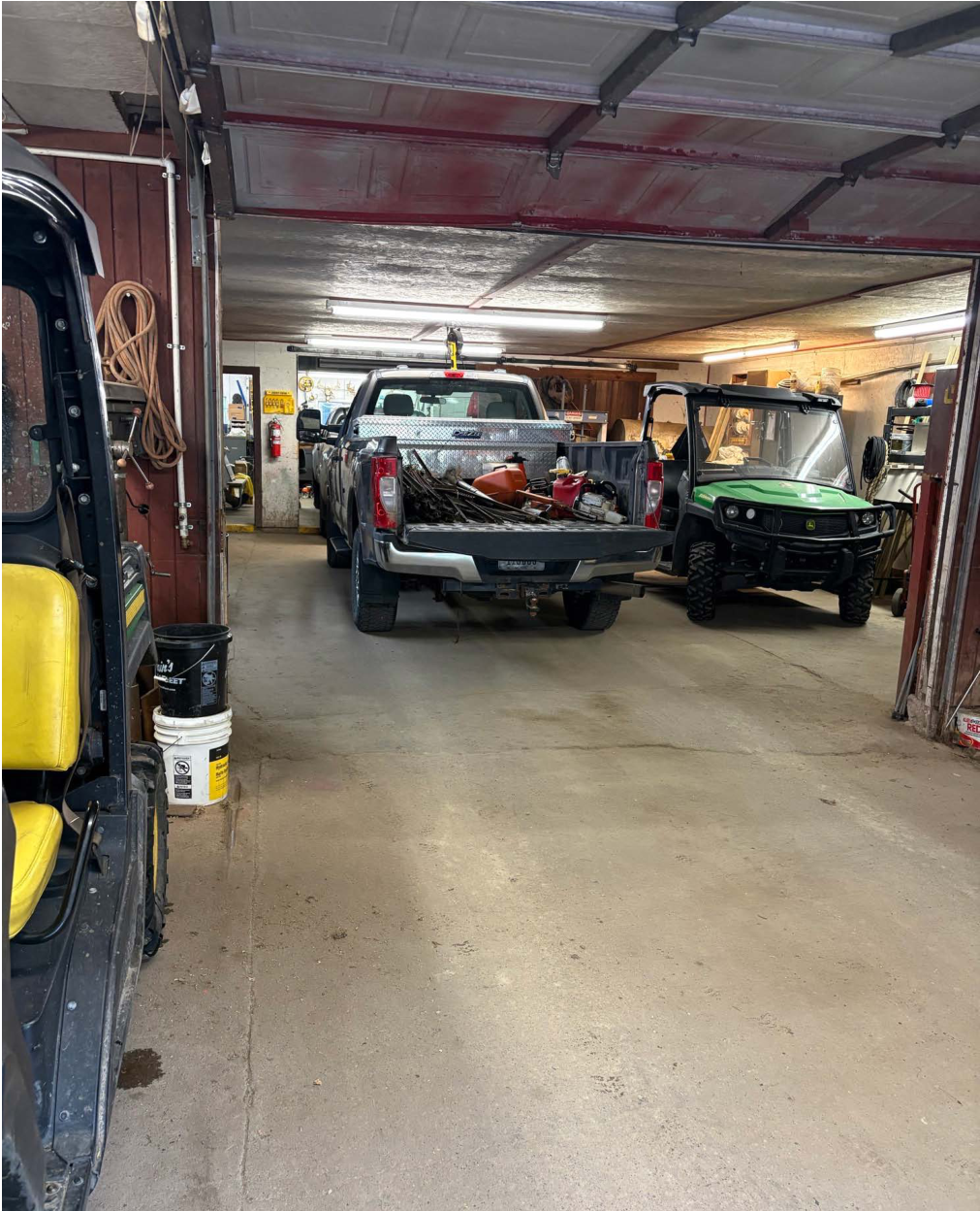 The image captures an interior garage space with two parked vehicles. A silver pickup truck occupies the center, its bed filled with tools and equipment, while a green utility vehicle is parked to the right. The garage has overhead lighting and wooden paneling, with scattered tools and equipment visible in the surroundings. The layout indicates a functional working environment.