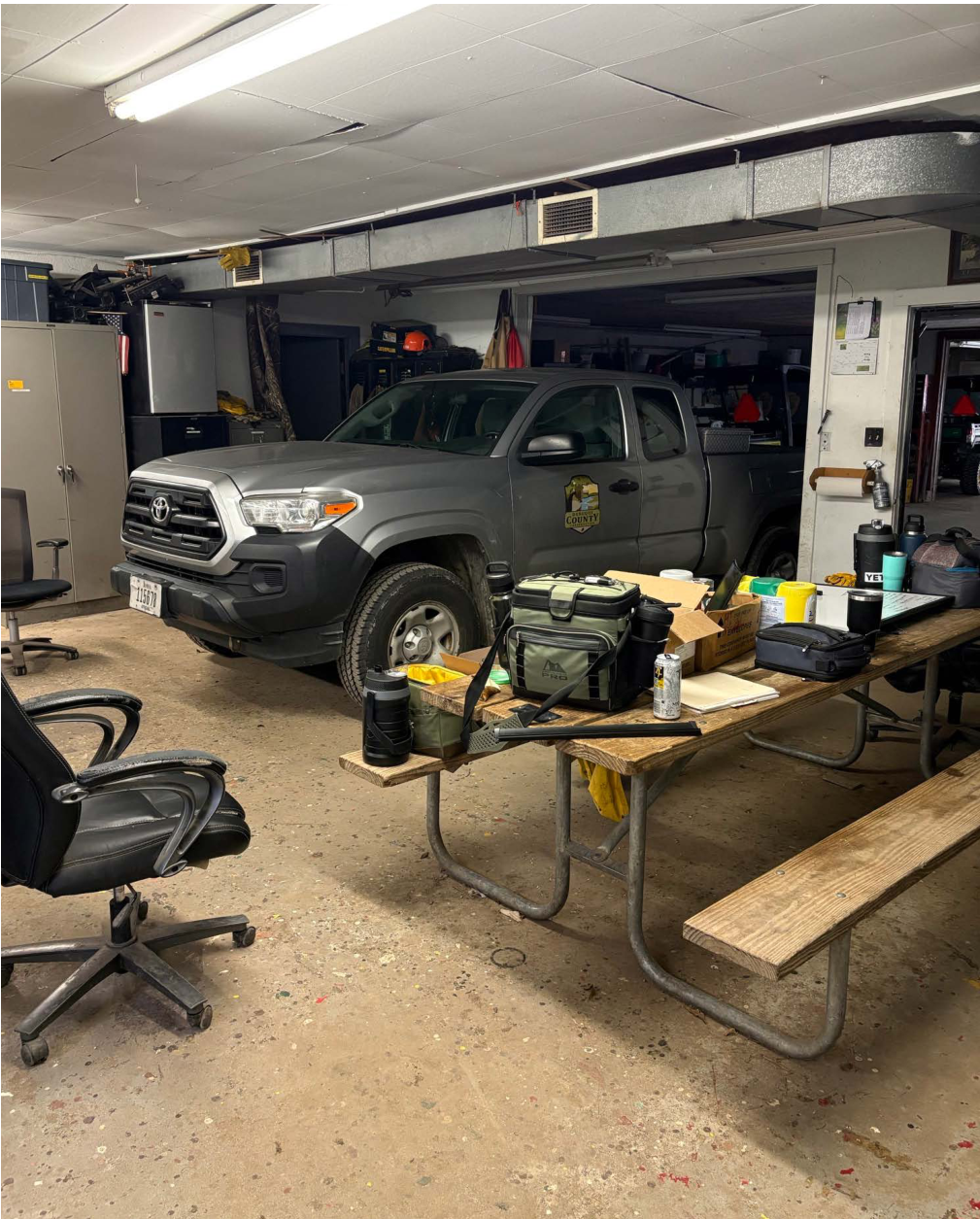The image depicts a garage space with a gray Toyota truck parked inside. In the foreground, there is a wooden picnic table cluttered with various items such as bags, beverages, and equipment. An office chair is seen in the left corner. The garage background includes storage units and tools, indicating a workspace environment.