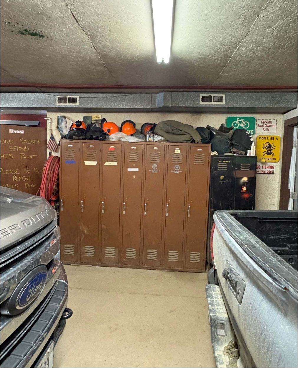 The image shows a locker area in a building. There are several brown metal lockers lined up against the wall. On top of the lockers, there are helmets, a jacket, and other safety gear. Visible notices and signs on the walls indicate various instructions and rules for the area, including notices about noise and fishing restrictions. The flooring appears to be concrete with vehicles parked nearby, indicating this might be a utility or maintenance space.