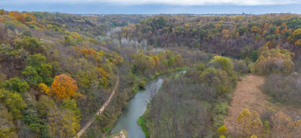 The image captures a scenic aerial view of a wooded area featuring a winding river flowing through it. The trees display a variety of fall colors, including orange, yellow, and green, highlighting the seasonal transition. Various patches of land are visible next to the river, with a mix of dense forest and open fields, contributing to the overall natural landscape.