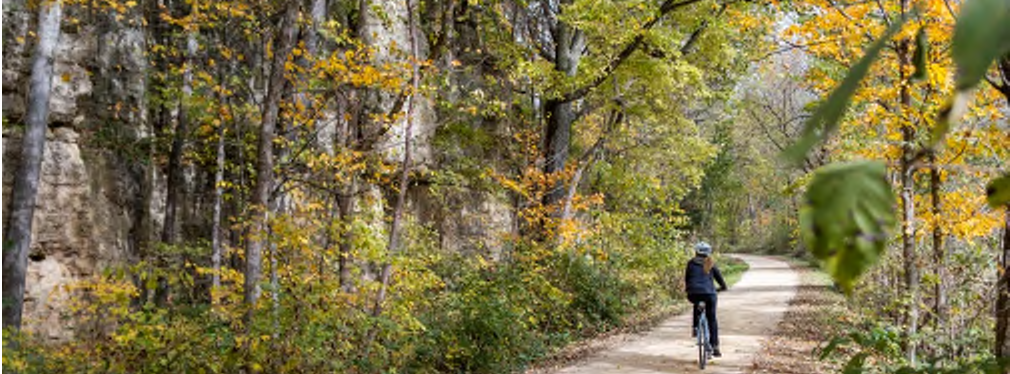 The image depicts a cyclist biking along a wooded trail. The pathway is flanked by trees with vibrant autumn colors, showcasing a mixture of greens and yellows. The terrain features a slight incline with rocky cliffs visible on one side, indicating a natural landscape. The cyclist is seen wearing a helmet and casual attire, illustrating an outdoor recreational activity.