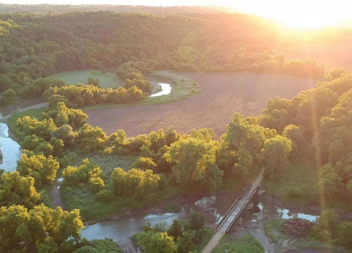 The image features an expansive aerial view showing a vibrant landscape with dense green trees surrounding a winding river. In the foreground, there is a narrow dirt road leading to a wooden bridge crossing over the river. Fields are visible on the landscape, giving a contrast to the greenery. The golden hue of the sunset bathes the entire scene, enhancing its natural beauty.