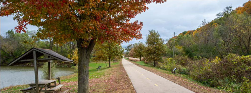 The image depicts a paved pathway winding along a riverbank, with benches and trees displaying vibrant autumn foliage on either side. The path is well-maintained and lined with grass, offering a peaceful setting for walking or biking. In the distance, there are signs indicating amenities and the continuation of the pathway. The overall atmosphere is tranquil, highlighting nature's beauty in the fall.