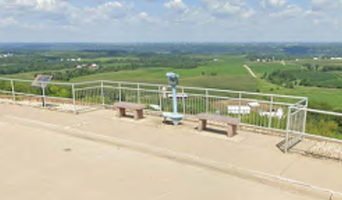 The image captures a viewpoint area equipped with two benches and a telescope positioned for observation. The surrounding landscape features lush green fields and distant hills under a partly cloudy sky. A small informational sign stands near the viewpoint, adding context to the scenery.