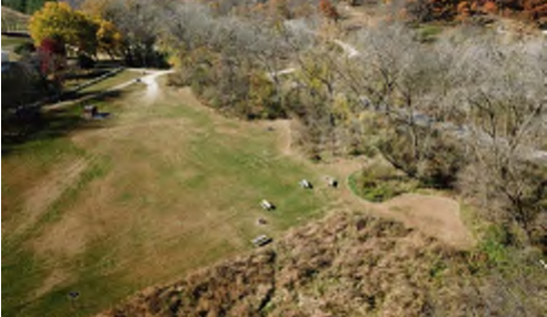 The image shows an aerial view of a park landscape featuring rolling grassy areas, clusters of trees, and clear winding paths that navigate through the terrain. The park appears to have several open spaces and a few benches scattered throughout the area, providing a serene natural environment.