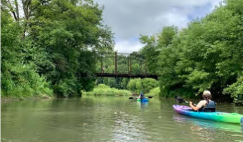 The image depicts a serene water scene with two individuals in kayaks, navigating the calm waters of a river. The kayakers are surrounded by lush green trees on both sides of the waterway. In the background, a distinctive, old iron bridge spans the river, adding a sense of depth and perspective to the scene. The sky is overcast, suggesting a moderately cloudy day.
