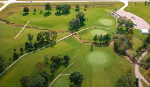 The image depicts a golf course layout from above, showcasing multiple green areas, fairways, and walking paths throughout the course. There are several circular greens, densely wooded areas, and a parking facility located at the top right of the image. The clear paths and layout provide an overview of the terrain and its spatial relationships.
