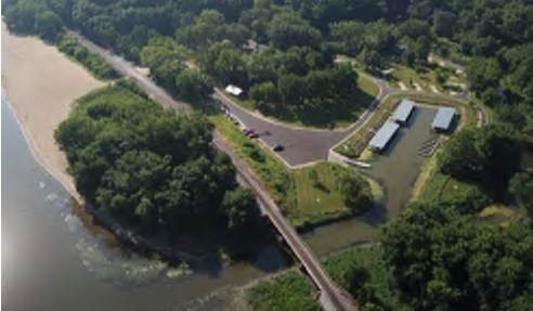 The image shows an aerial view of a park near a river. To the left, there is a sandy beach area. In the center, a parking lot accommodates several vehicles, leading to a series of structures possibly used for park services. The area is surrounded by greenery, indicating wooded regions that provide a natural habitat. The spatial arrangement includes the river forming one boundary of the park and the beach providing access to water activities.