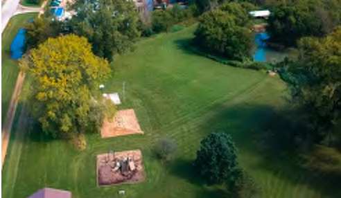 The image shows an aerial view of a large grassy area marked by a few trees, two small dirt patches, and a creek flowing along one side of the space. The natural layout is open with a mix of greenery, providing a sense of the land's openness and topography.