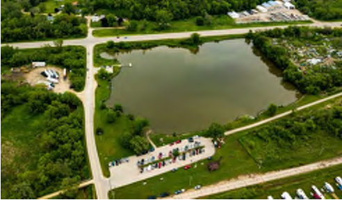 The image showcases an aerial perspective of a landscape featuring a small lake. Surrounding the lake are areas of greenery and a visible road network that includes intersections. A parking area is located on the bottom left, with several parked vehicles, while the upper part of the image shows additional buildings and areas of vegetation. The layout highlights the relationship between the parking, roadways, and natural features in the vicinity.