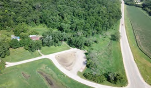 The image shows a birds-eye view of a rural landscape featuring a road that runs along the right side. A circular clearing is visible at the bottom center of the image, with a house and trees to the left, indicating a wooded area. The clearing appears to be under construction or excavation, while the surrounding area includes grassy fields and tree cover. The spatial layout indicates a transition from natural forested land to developed clearing and roadway.