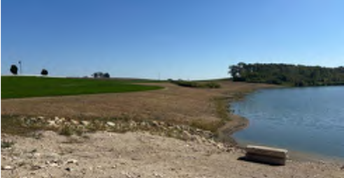 The image captures a tranquil lakeside scene with a sandy shoreline. On the left, there is a well-maintained grassy area with a few trees in the background. The water appears clear and calm, and the sky is bright blue, indicating a sunny day. The sandy beach in the foreground transitions to the grass further back, providing a natural setting for recreation or relaxation.