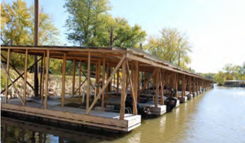 The image depicts a wooden dock with a series of supports extending over a calm body of water. The dock appears to have space for multiple boats and is framed by lush greenery on either side, indicating a tranquil outdoor setting. No specific text or labels are visible in the image.