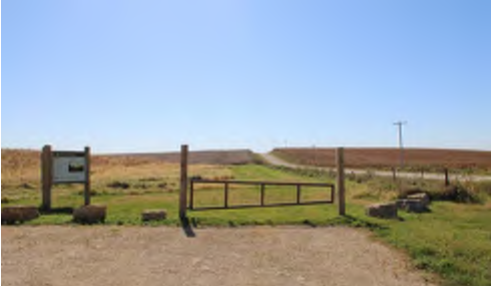 The image shows a wooden entrance gate located at the edge of a grassy area. Beyond the gate lies a dirt road that stretches out into the distant landscape, framed by fields under a clear blue sky. A sign is positioned next to the gate, providing information about the area, but the visible text on the sign is not clear.