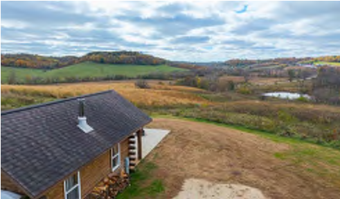 The image captures a scenic view from an elevated perspective, showcasing a house in the foreground with a sloped roof and wooden exterior. Surrounding the house, there are expansive fields with varying shades of green and brown, indicative of autumn. In the background, rolling hills create a picturesque landscape, and a small pond is visible amidst the terrain.