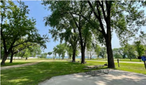The image depicts a park area with lush green grass and numerous tall trees. The sunlight filters through the leaves, creating a pleasant atmosphere. In the background, there is a glimpse of a body of water, suggesting proximity to a lake or river. A concrete pathway is visible, leading through the park.