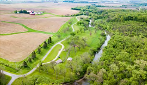 The image captures an aerial view of a park showcasing winding pathways, a creek flowing through the area, and scattered trees. In the background, there are fields alongside signs of agricultural activity. The arrangement of paths indicates routes for exploration, and the vegetation provides a natural setting, contributing to the park’s ambiance.