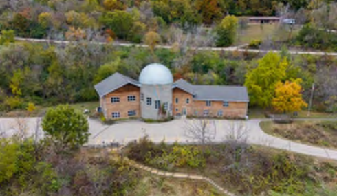 This image depicts an aerial view of a wooden structure featuring a prominent dome on its roof. The building is situated amidst a natural landscape filled with trees displaying autumn colors. A winding path leads up to the front of the building, surrounded by vegetation, which creates a serene setting. The paths are not labeled, but they appear to provide access to the building from various directions.