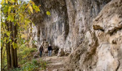 The image depicts a narrow trail alongside a rocky cliff. Two hikers can be seen walking on the path, with lush greenery and autumn foliage framing the scene. The cliff appears rugged, composed of exposed rock, while the trees on either side display a mix of green and yellow leaves, indicative of the fall season.