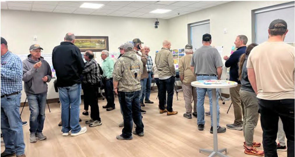 The image shows a gathering of individuals in a well-lit meeting room. Many attendees are standing and engaged in conversation, while others interact with presentation boards displaying various visuals. The room has a neutral-colored flooring and is furnished with tables. The atmosphere suggests a collaborative discussion or presentation session.